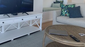 Una sala de estar con un televisor de pantalla plana sobre un mueble blanco, una mesa de centro de mimbre con una computadora portátil y un control remoto, un sofá gris con cojines estampados y una ventana con un valance a rayas.