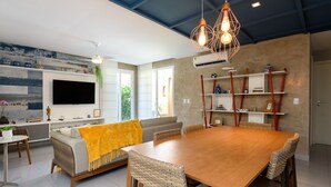 Ein modernes Wohnzimmer mit einem Holzesstisch, Korbstühlen, einem Sofa mit gelber Decke, einem flachbildfernseher an der Wand und einem Bücherregal mit roten Akzenten.
