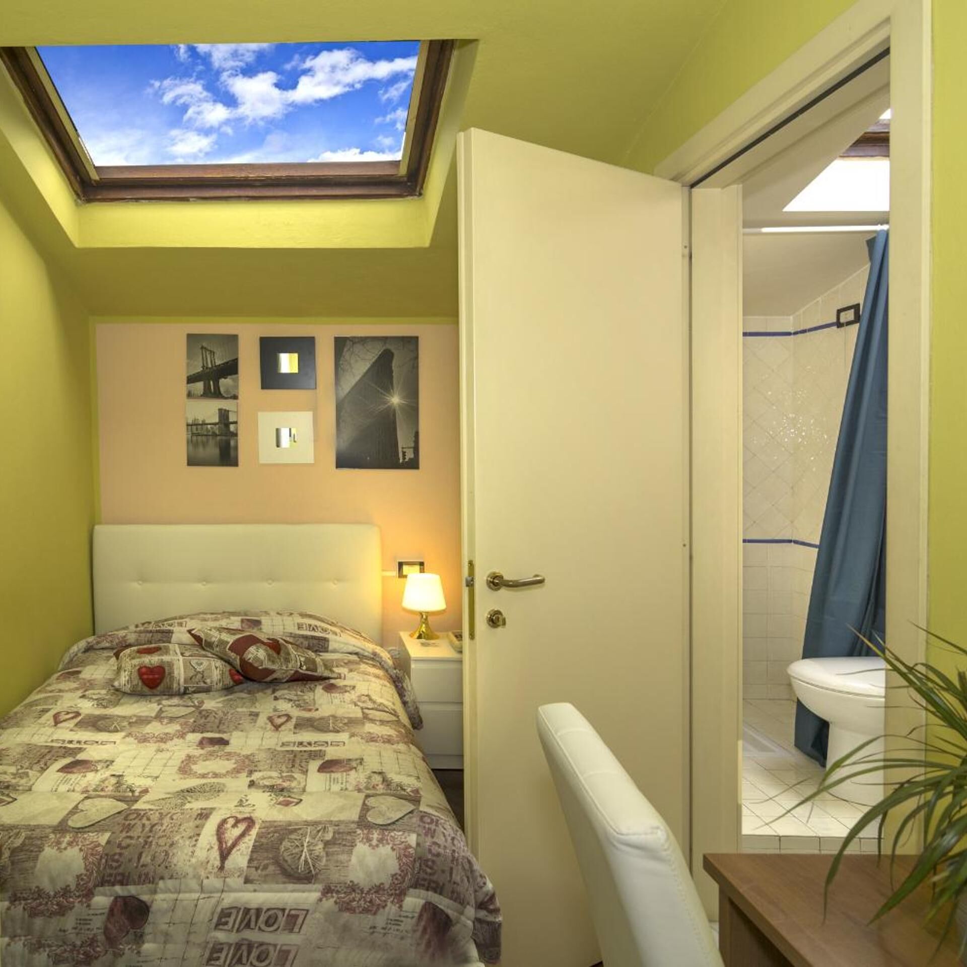 A bedroom with a skylight, a bed with a patterned bedspread, a nightstand with a lamp, and a bathroom visible through an open door.