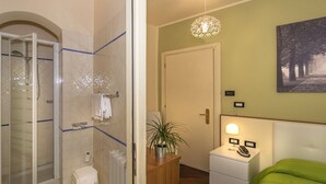 A bathroom with a shower, toilet, and a sink. There is a small bedside table with a lamp and a phone. A green bed is visible in the corner. - Hotel Il Giardino (Pisa)