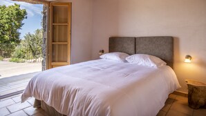 Et soveværelse med en stor seng, et træsengegærde og et natbord med en lampe. En trædør fører til et udendørs område med udsigt over træer og en balkon.