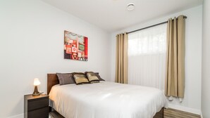 Une chambre à coucher avec un lit, une table de chevet, une lampe, un tableau et une fenêtre avec des rideaux.
