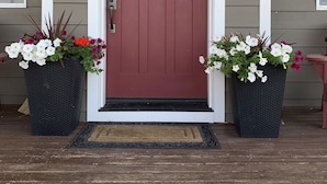 Puerta roja con un adorno de corona, dos macetas con flores y un felpudo.