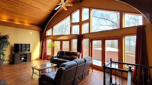 A spacious living room with a leather sofa, a glass coffee table, and a flat-screen TV. Large windows provide ample natural light and a view of the outdoors.