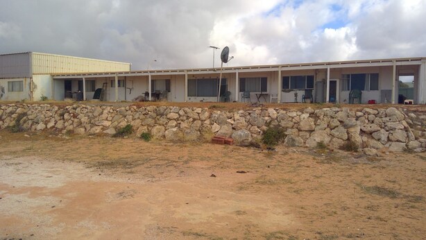 Un edificio de techo plano, con varias ventanas y una antena parabólica.  Una pared de piedra se encuentra frente al edificio.