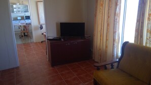 Una sala de estar con televisor sobre un mueble de madera, un sofá y una cocina al fondo.