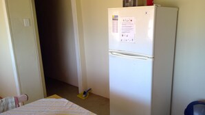 Una habitación pequeña con un refrigerador blanco, un utensilio de limpieza amarillo y una cama con colcha a rayas.