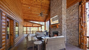Una cabaña de troncos con terraza de madera, muebles de mimbre, chimenea de piedra y un letrero que dice 