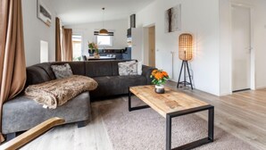 A modern living room with a grey sectional sofa, a wooden coffee table, and a floor lamp. A kitchen is visible in the background.