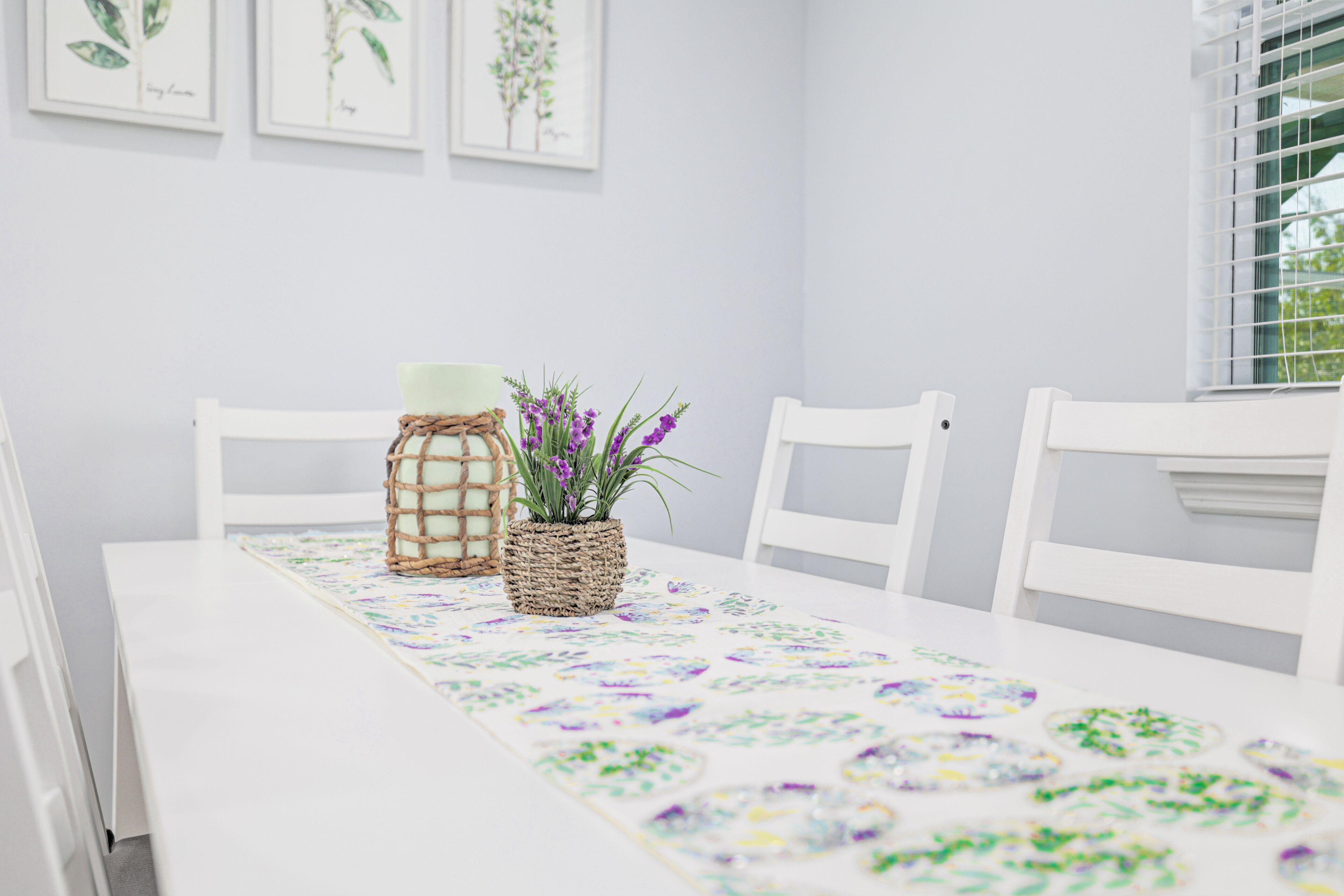 Una mesa blanca con camino de mesa floral, dos cestas de mimbre y una maceta con flores moradas.