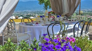 An outdoor dining area with a table set for a meal, surrounded by white chairs, and overlooking a scenic view of a city and mountains. - Villa with Pool in Tuscany for 8 People (Cantagrillo)
