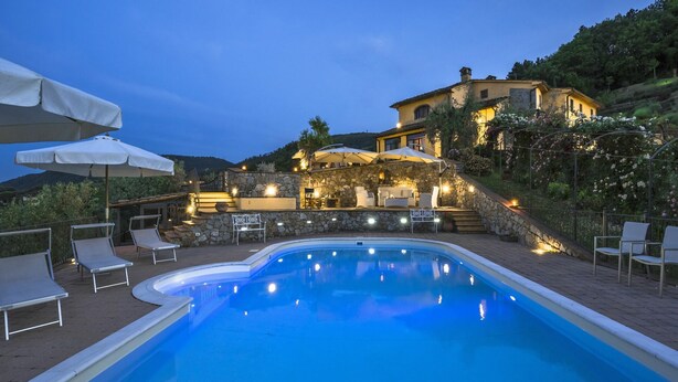 A swimming pool with lounge chairs and umbrellas, a stone wall, and a building with outdoor seating and lights in the background. - Villa with Pool in Tuscany for 8 People (Cantagrillo)