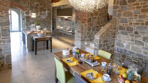 Cozinha rústica com paredes de pedra, uma mesa de jantar de madeira posta para o café da manhã e um lustre moderno.