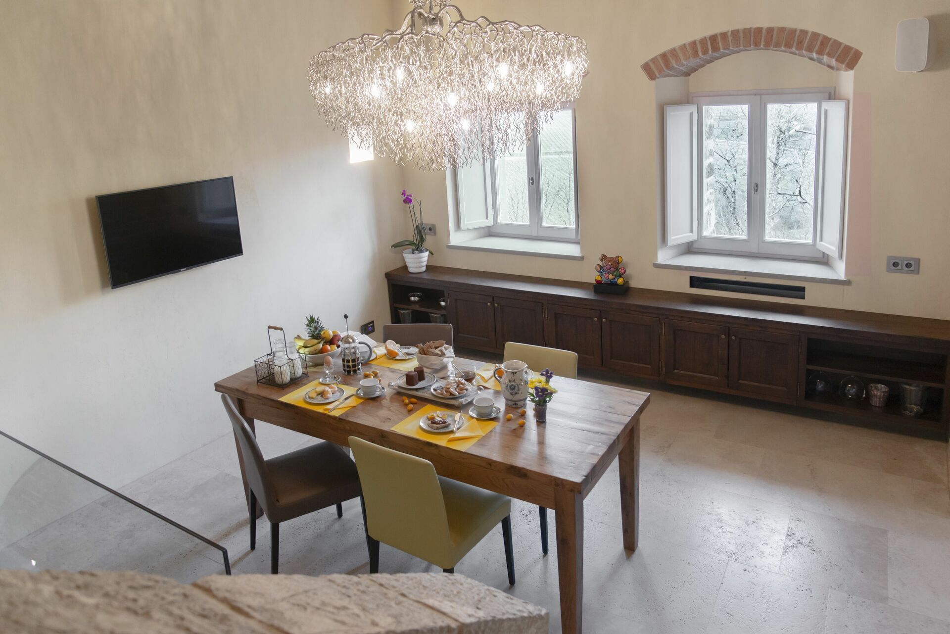 Un coin repas avec une table en bois dressée pour le petit-déjeuner, un lustre et une cuisine en arrière-plan.