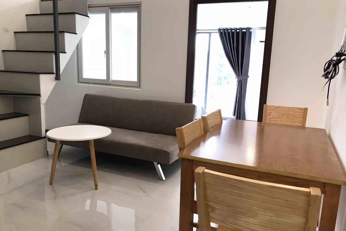 Uma sala de estar moderna com um sofá, uma mesa redonda e cadeiras de madeira. Uma escada é visível à esquerda e uma janela com cortinas à direita.