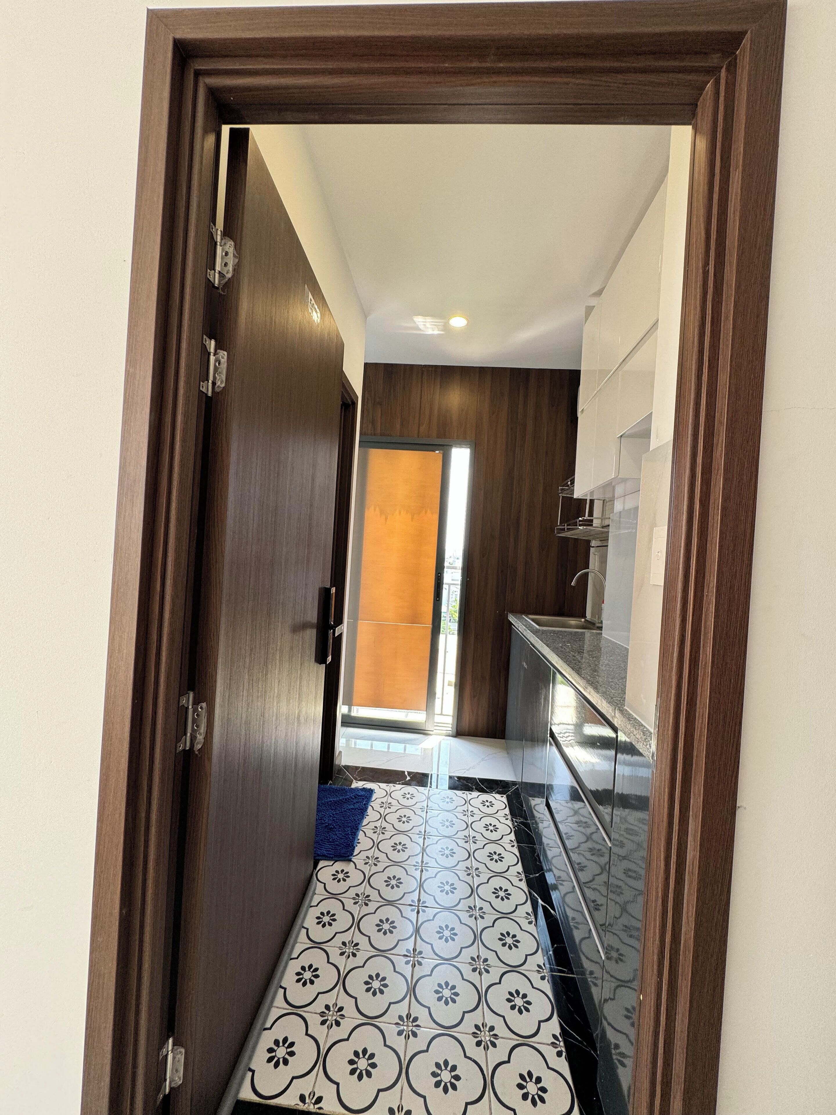 A hallway with a wooden door, patterned floor tiles, and a view into a room with a window and a door.