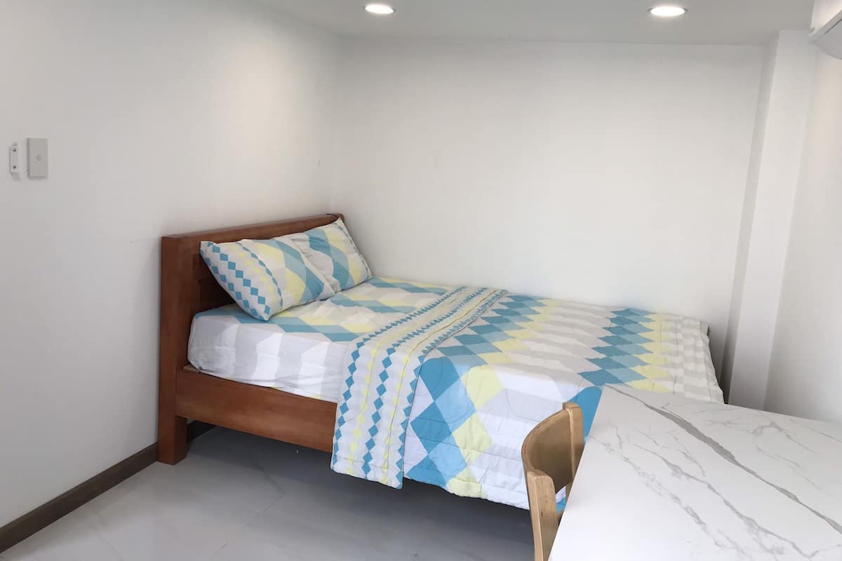 Uma cama de solteiro com cabeceira de madeira, uma colcha estampada e uma mesa de mármore num quarto com paredes e luminárias de teto brancas.