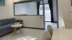 Un salon moderne avec un canapé gris, une table ronde agrémentée d’une plante et une grande fenêtre avec des rideaux.