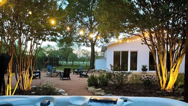 A hot tub with a waterfall feature, surrounded by trees and string lights, with a patio area and a white building in the background.