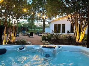 Un bain à remous doté d’une cascade, entouré d’arbres et illuminé de guirlandes lumineuses, avec un espace patio et un bâtiment blanc en arrière-plan.