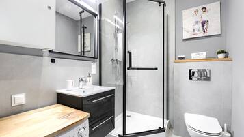 Un baño moderno con cabina de ducha de vidrio, inodoro blanco, tocador de madera y estante con artículos de aseo.
