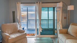 Una stanza con due divani beige, un'ampia porta scorrevole in vetro che dà su un balcone con vista sulla spiaggia e una lampada da terra.