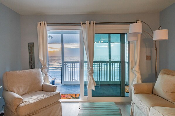 Um quarto com dois sofás bege, uma grande porta de correr de vidro que dá para uma varanda com vista para a praia, e um abajur.