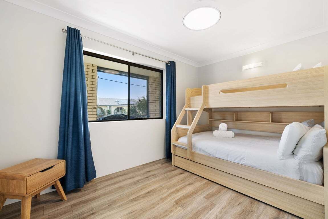 Un dormitorio con una cama individual superior e inferior, una mesita de madera, una ventana con cortinas y vistas a edificios.