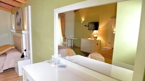 A hotel room with a large mirror, a bed, a bedside table with a lamp, a small table with a chair, and a television on the wall. - White City (Berat)