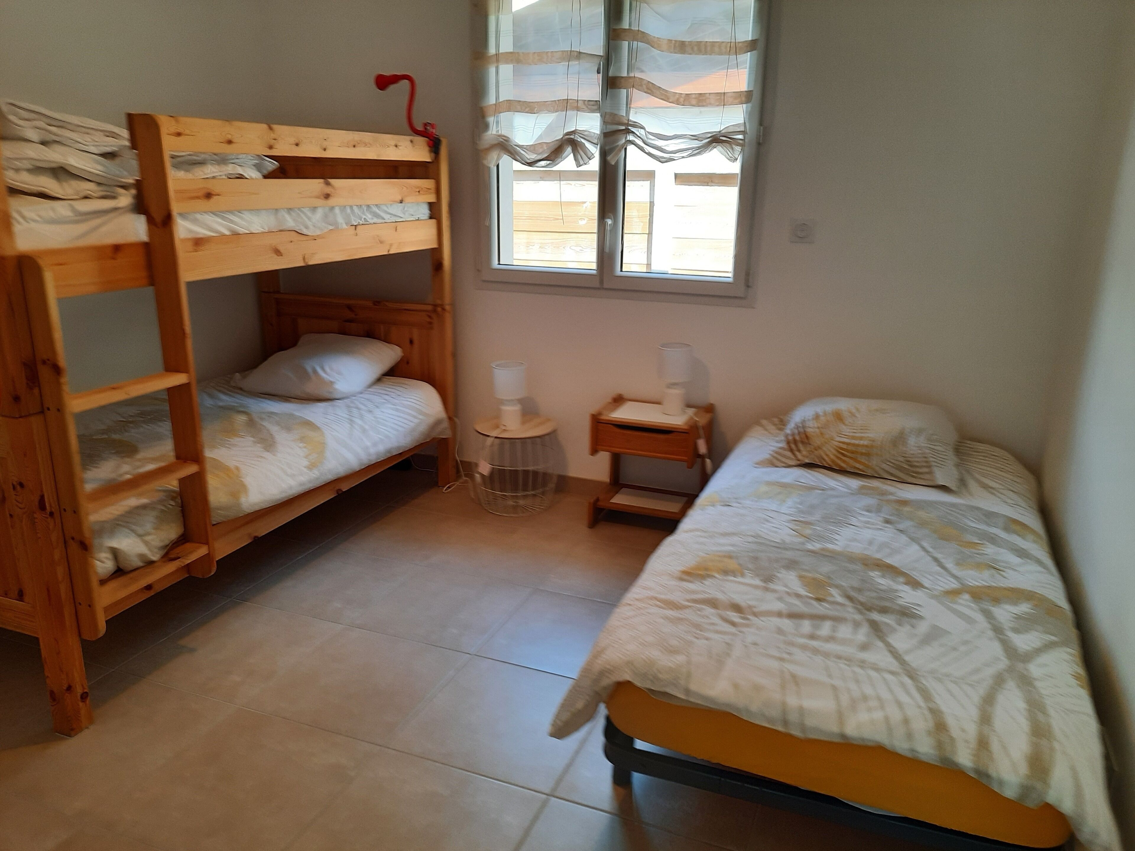 A dormitory room with two bunk beds, a small bedside table with a lamp, and a window with sheer curtains.