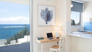 A modern room with a view of the ocean, a desk with a laptop, a chair, and a framed artwork on the wall.