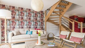Ein Wohnzimmer mit Sofa, Couchtisch und Sesseln, eine Treppe und eine Küche im Hintergrund.