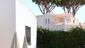 A pool area with lounge chairs and umbrellas, a white building, and a hedge with trees. - Casa Azevedo - Vilamoura (Vilamoura)
