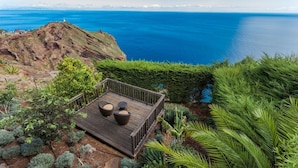 Une terrasse en bois avec deux fauteuils en osier et une table, offrant une vue sur un paysage côtier avec une falaise rocheuse et l’océan.