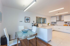 Uma cozinha moderna com armários brancos, uma mesa de jantar de vidro e uma janela com persianas.