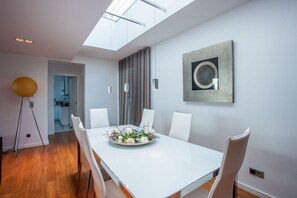 Uma sala de jantar moderna com mesa branca, cadeiras brancas, uma janela grande e uma obra de arte contemporânea na parede.