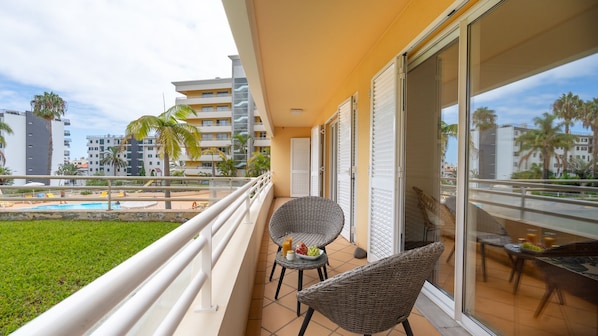 Un balcone con sedie in vimini, un tavolino con della frutta e vista su una piscina e degli edifici.