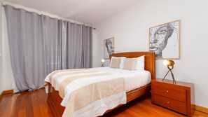 A bedroom with a wooden bed, grey curtains, a bedside table with a lamp, and two wall art pieces.