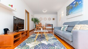 A living room with a TV, wooden entertainment unit, a grey sofa, a patterned rug, and a painting of a beach scene.
