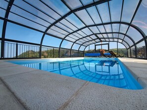 Piscina con tetto in vetro, circondata da una ringhiera in metallo, con vista su un paesaggio collinare.