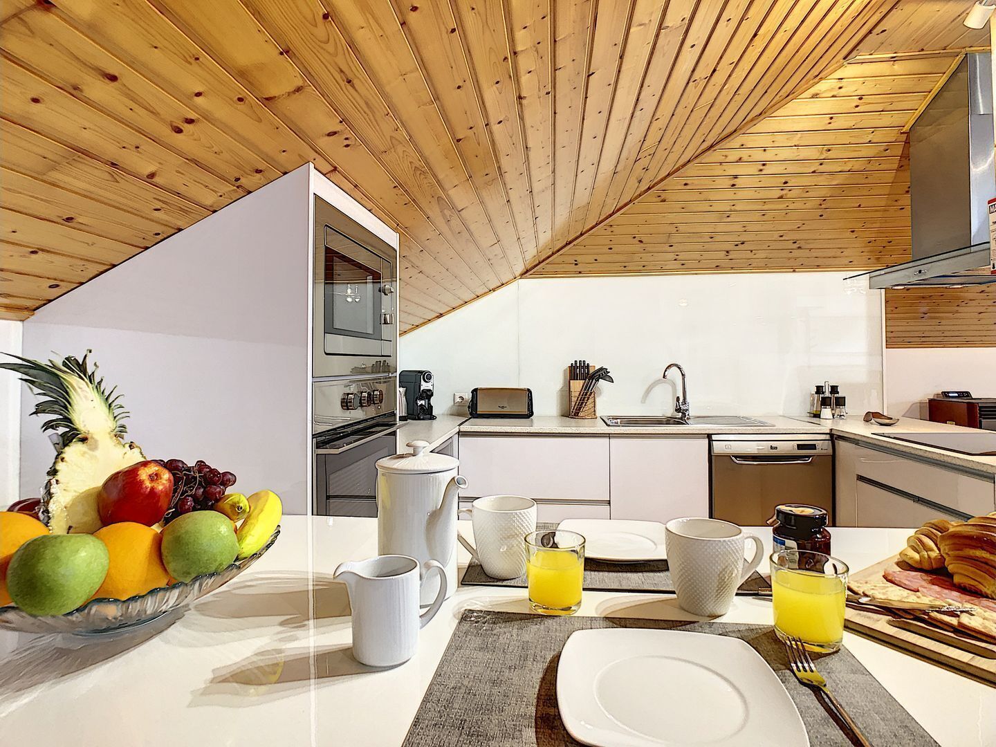 Een moderne keuken met houten balken in het plafond, roestvrijstalen apparatuur en een ontbijtgedeelte met fruit, koffie en croissants.