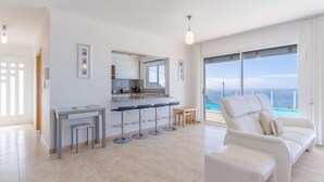 Een moderne woonkamer met een witte bank, een eethoek met barkrukken, een keuken met een groot raam en uitzicht op de oceaan.