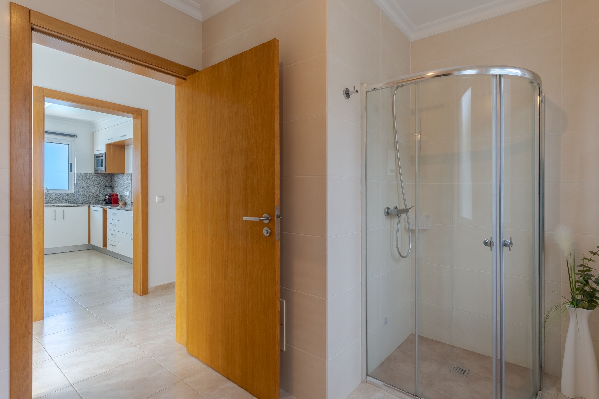 A modern bathroom with a glass shower enclosure and a wooden door leading to a kitchen area.
