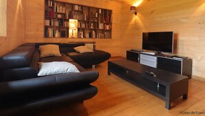 Een gezellige woonkamer met een zwarte leren bank, een houten boekenkast, een flatscreen televisie en een salontafel.