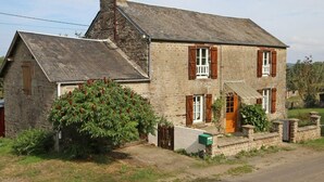 A stone house with a sloped roof, wooden shutters, and a green bush in front.