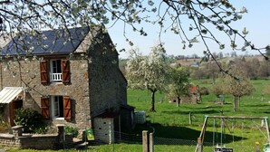 A stone house with a balcony, surrounded by a rural landscape with green fields and trees.