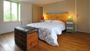 A bedroom with a wooden floor, a large bed with white bedding, a wooden headboard, a nightstand with a lamp, a chest of drawers, and a window with a view of trees.
