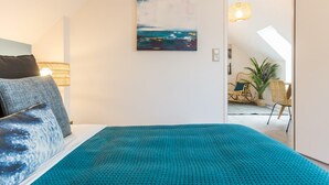 A bedroom with a bed, pillows, a painting, and a view into a living area with a chair and a plant. - Independent house in Portbail, pets allowed, near the beach (Portbail)