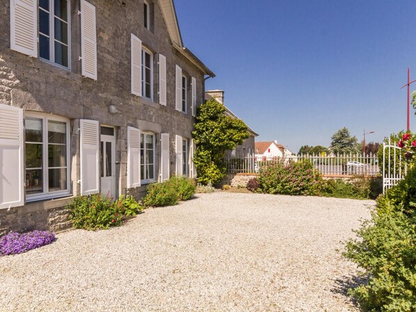 A stone building with multiple windows and shutters, a gravel driveway, and a fenced area with greenery and flowers.