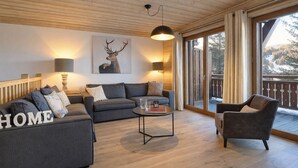 Ein modernes Wohnzimmer mit einer Sofagarnitur, einem Couchtisch und einem Sessel. An der Wand hängt ein Bild eines Rehs, und der Raum hat eine Holzbalkendecke und große Fenster.